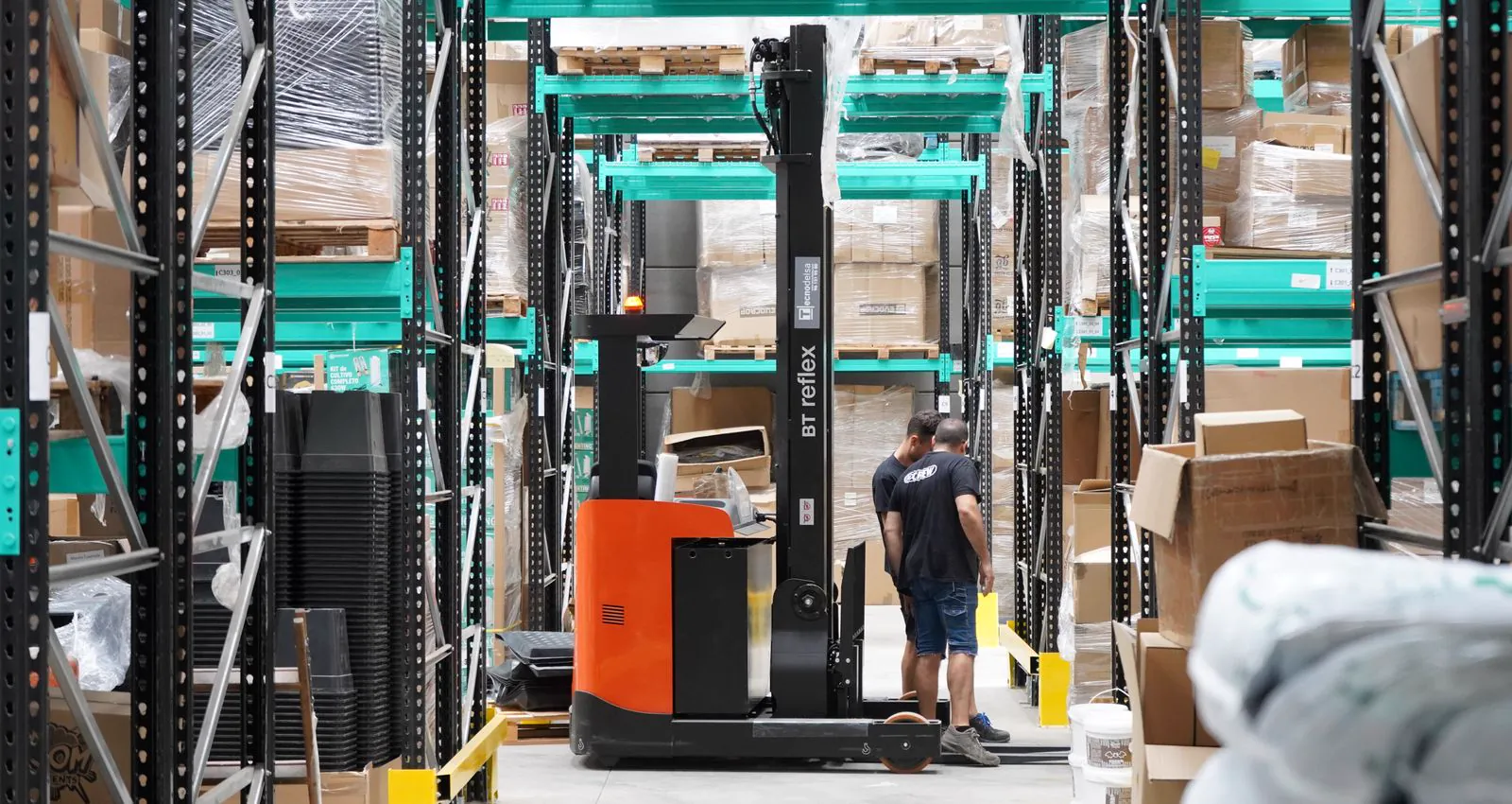 Warehouse staff working a forklift between racked pallet aisles